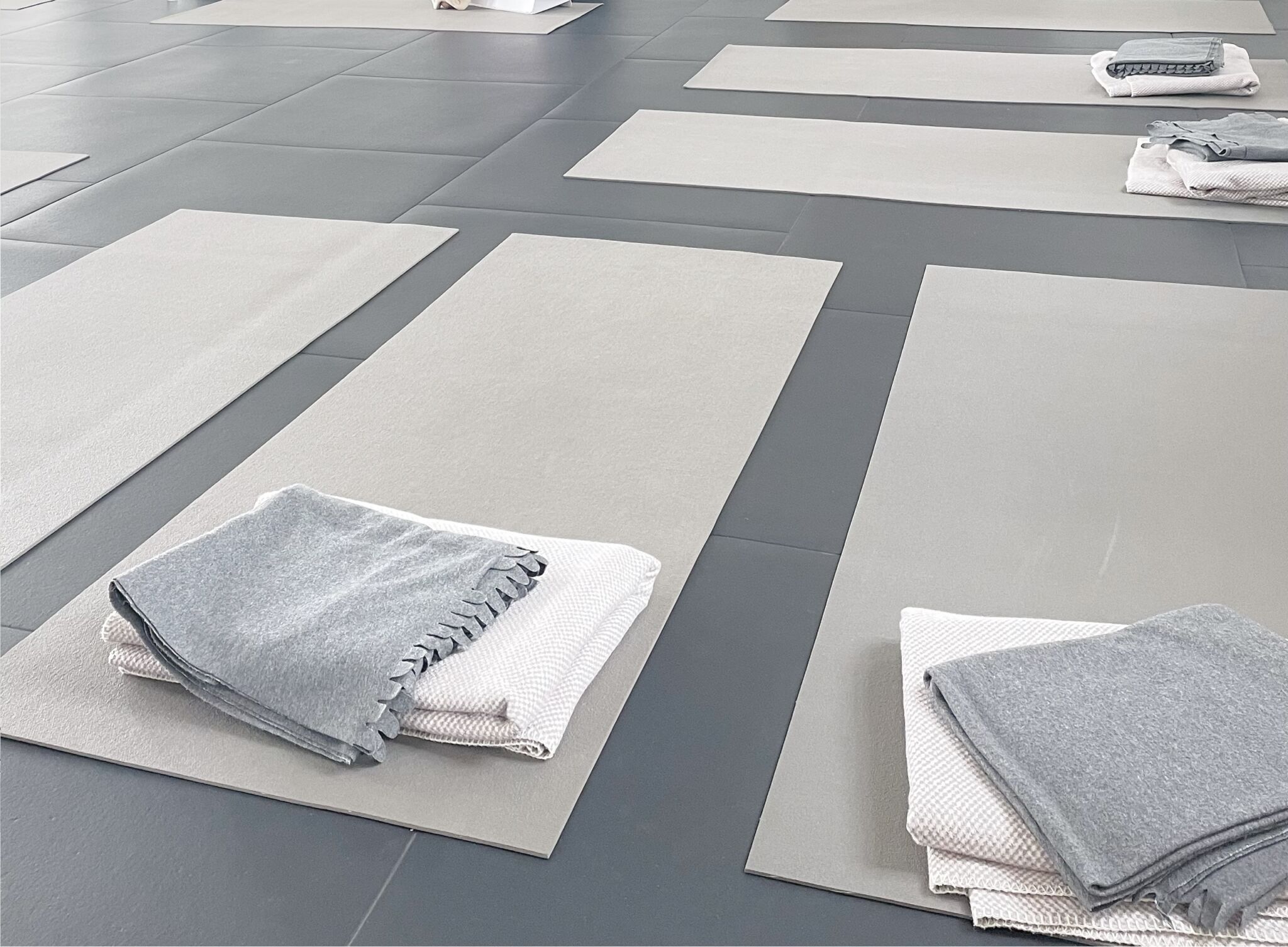 Yogamåtter ligger på et flisebelagt gulv med tæpper pænt foldet ovenpå hver måtte, hvilket indikerer forberedelse til en yogaklasse i et roligt studie.