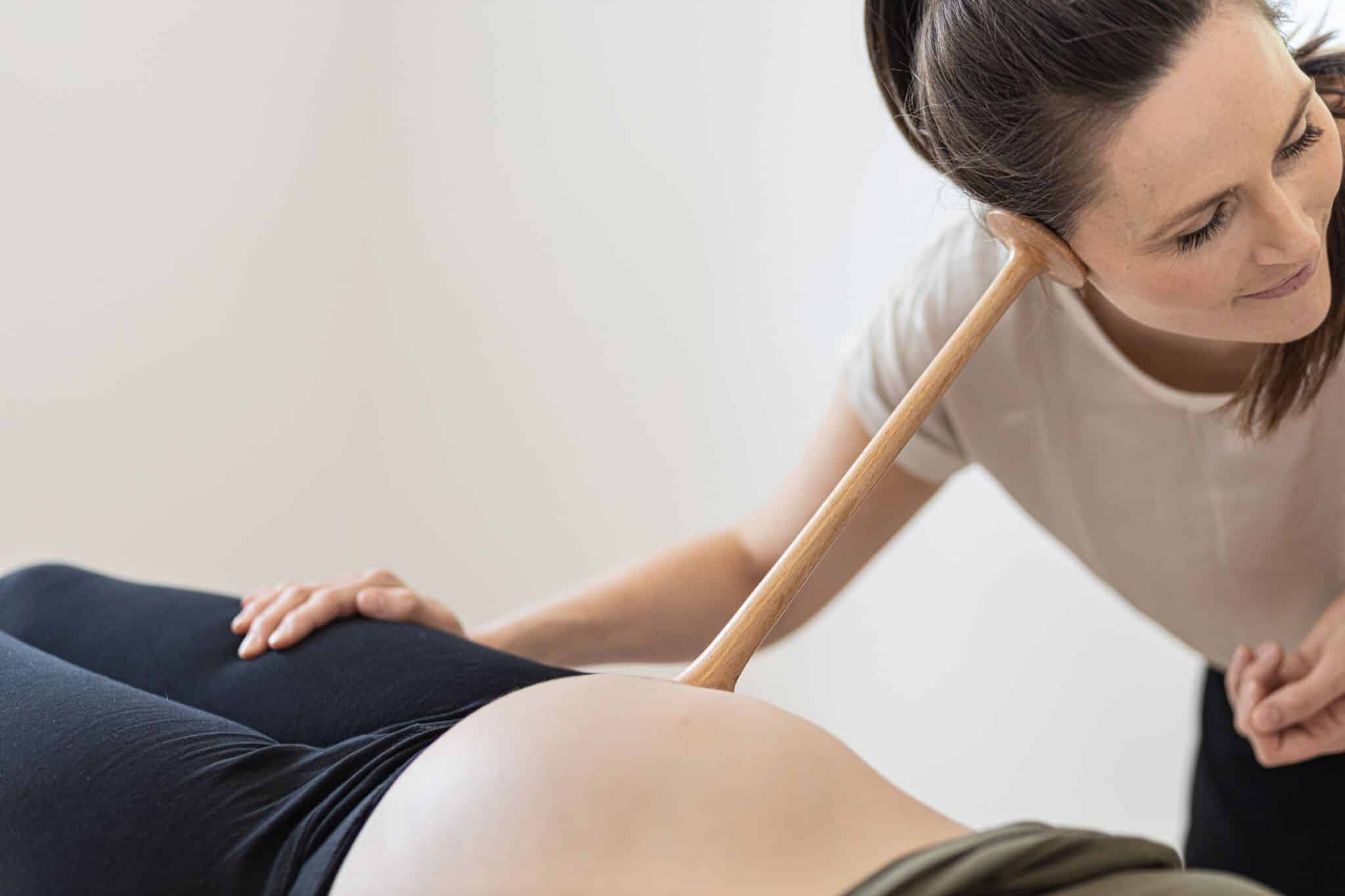 Kvinde lytter med træstetoskop til gravid mave i lys, minimalistisk rum. Hun smiler opmærksomt, mens hendes hånd hviler på patientens ben.