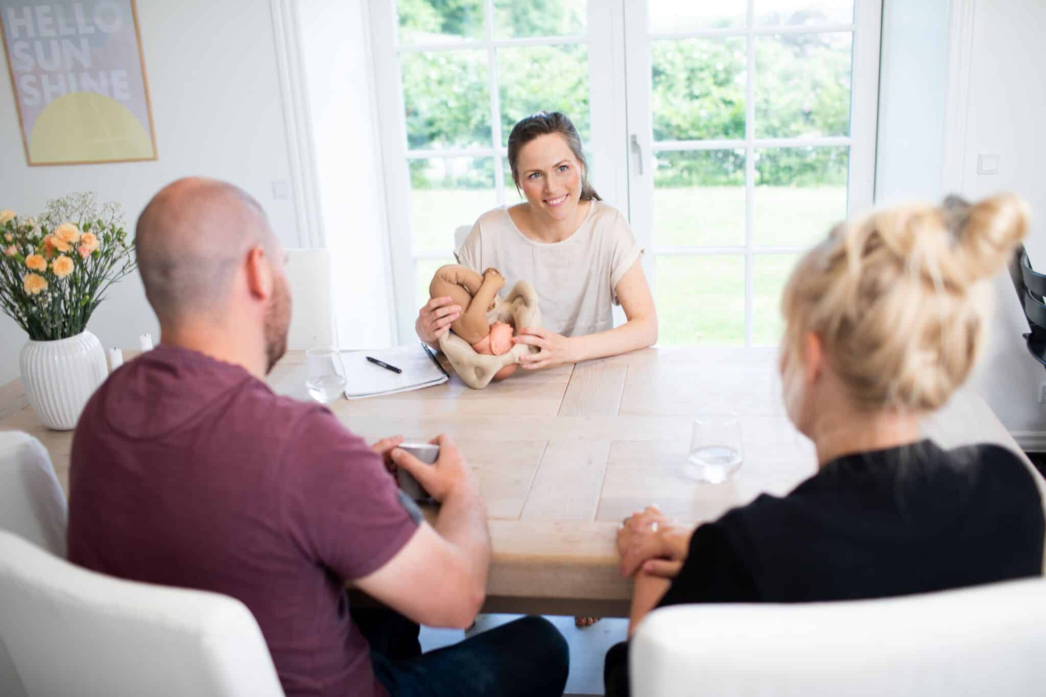 Kvinde holder en model af et foster under en samtale med to personer ved et træbord. Væggene har en plakat med teksten “HELLO SUNSHINE” og en vase med blomster.