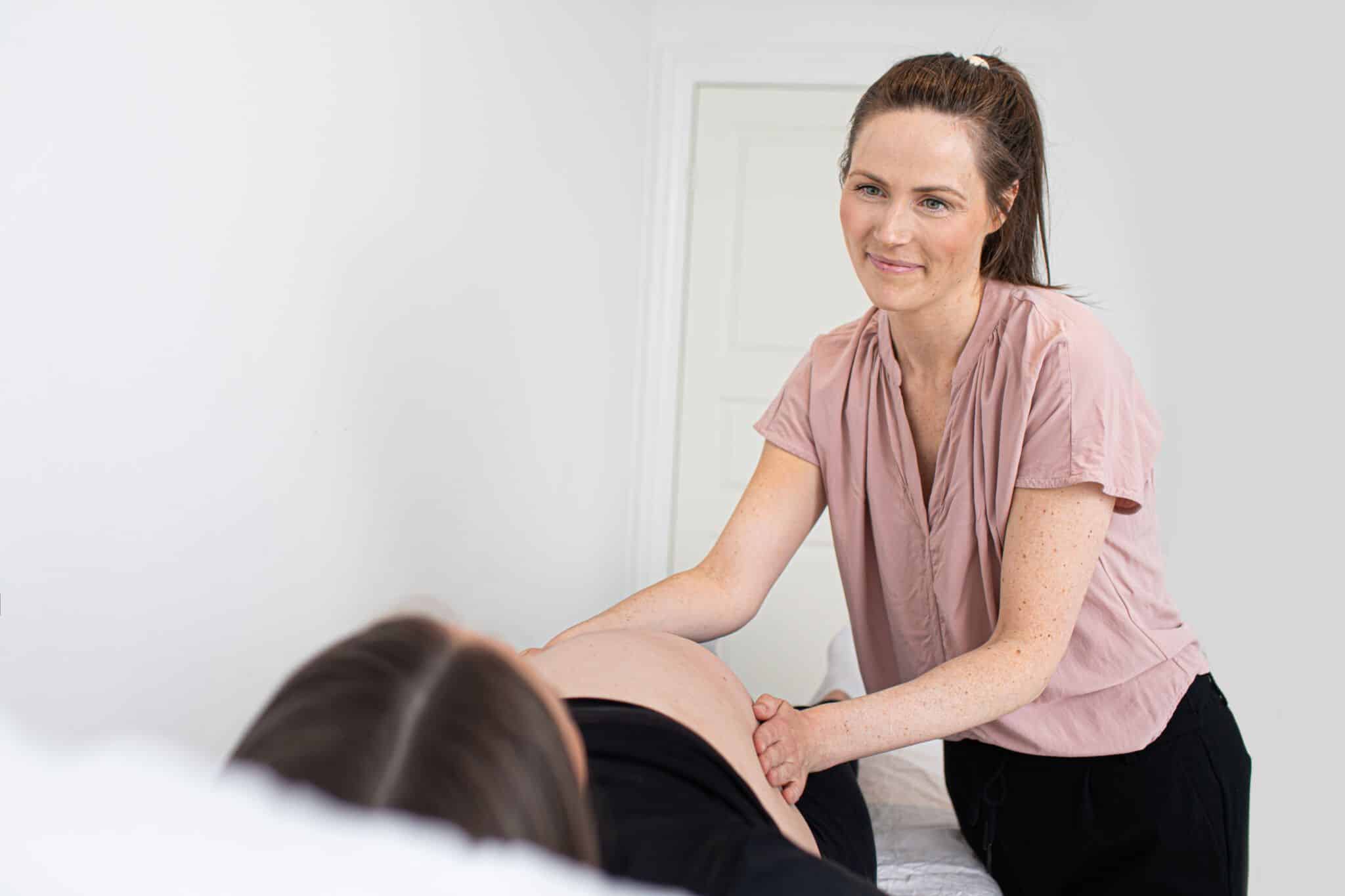 En kvinde masserer en gravid mave i et lyst rum med hvid baggrund. Den gravide ligger ned, mens den anden kvinde smiler blidt.